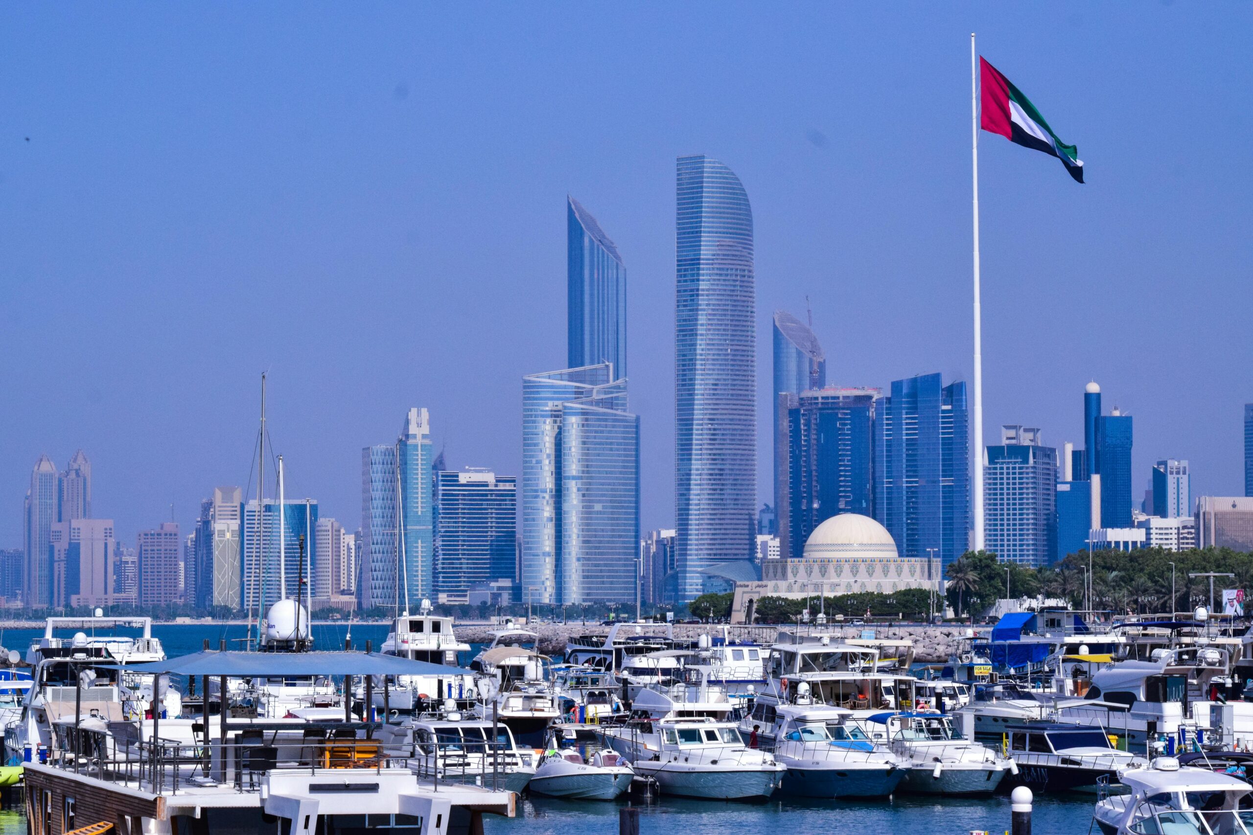 Services A scenic view of Abu Dhabi marina with modern skyscrapers and a flag in the foreground.